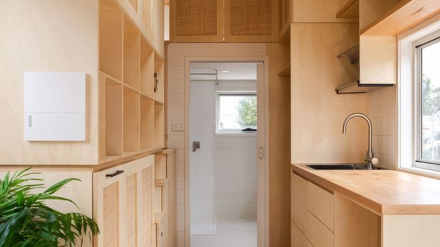 A compact, warm-toned kitchen and utility area with extensive light timber cabinetry, open shelving, integrated storage, a timber benchtop and stainless-steel sink. A bright tiled bathroom or utility space is visible through the doorway, while a white appliance and indoor plant sit to the left. The cabinetry includes woven or perforated panel details and the room is illuminated by a large side window.
