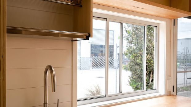 Bright interior view of a contemporary kitchen, focused on a stainless-steel gooseneck tap, timber benchtop, timber cabinetry, and a large sliding window with views to the exterior. Horizontal wall panelling and exposed timber ceiling beams add warmth to the space.