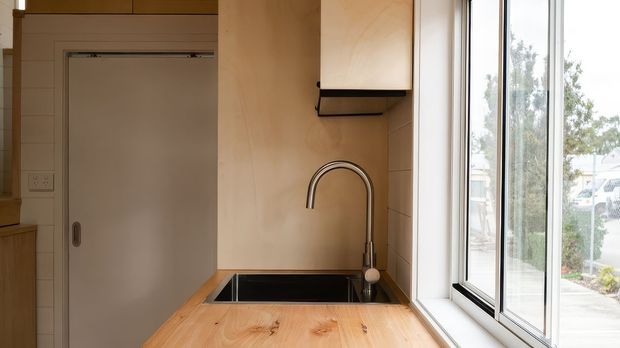 Compact kitchen or kitchenette viewed at the sink, with a warm timber benchtop, dark rectangular undermount sink, curved chrome tap, pale timber wall cabinetry, and a large black-framed window. A plain internal door and additional cabinetry are visible to the left. The space is simply detailed, warm, and naturally lit.