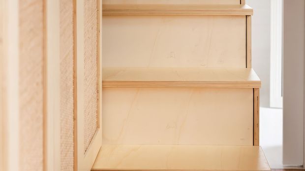 Close-up of custom light timber built-in joinery, showing a stepped shelving or stair-like arrangement with exposed plywood edges and smooth pale wood panels. A white-framed window is partially visible at the right edge. The image focuses on the craftsmanship and material detailing rather than a full room.