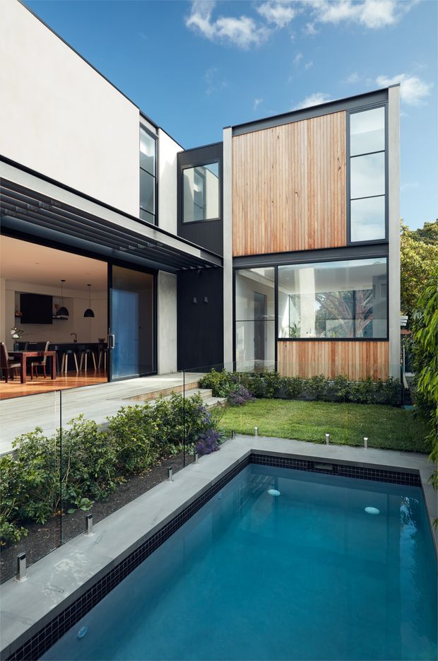 Contemporary multi-level courtyard residence viewed from the outdoor pool area. The house combines crisp white and dark charcoal rendered walls with extensive glazing and a prominent vertical timber-clad upper facade. An open-plan kitchen and dining area extends onto a pale deck, while a landscaped garden borders the rectangular swimming pool in the foreground.