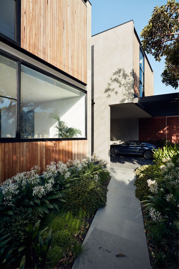 A contemporary multi-level residential facade viewed from a landscaped front walkway. The building combines vertical timber cladding, smooth pale rendered walls, dark metal-framed glazing, and a projecting carport canopy with a brick wall behind it. Dense layered planting borders a large-format grey paved path leading toward the sheltered driveway and parked car. Strong sunlight creates crisp shadows across the facade and garden.