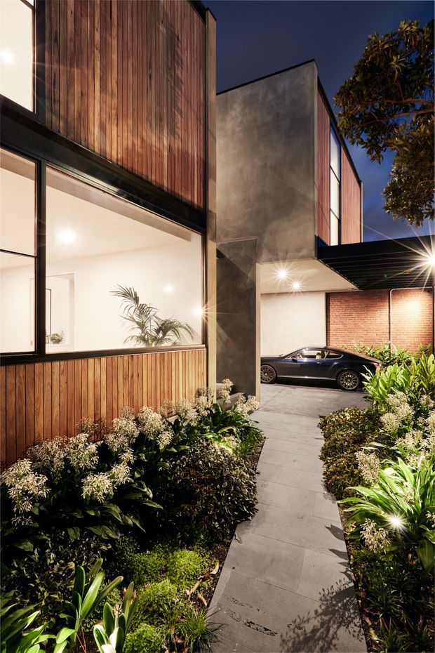 Night-time exterior view of a contemporary multi-level residence with warm vertical timber cladding, large black-framed glazed openings, a raw concrete feature wall, and a covered carport. A paved walkway passes through dense landscaped planting toward the illuminated entrance and parked car. Red brickwork and strong architectural lighting add texture and contrast.