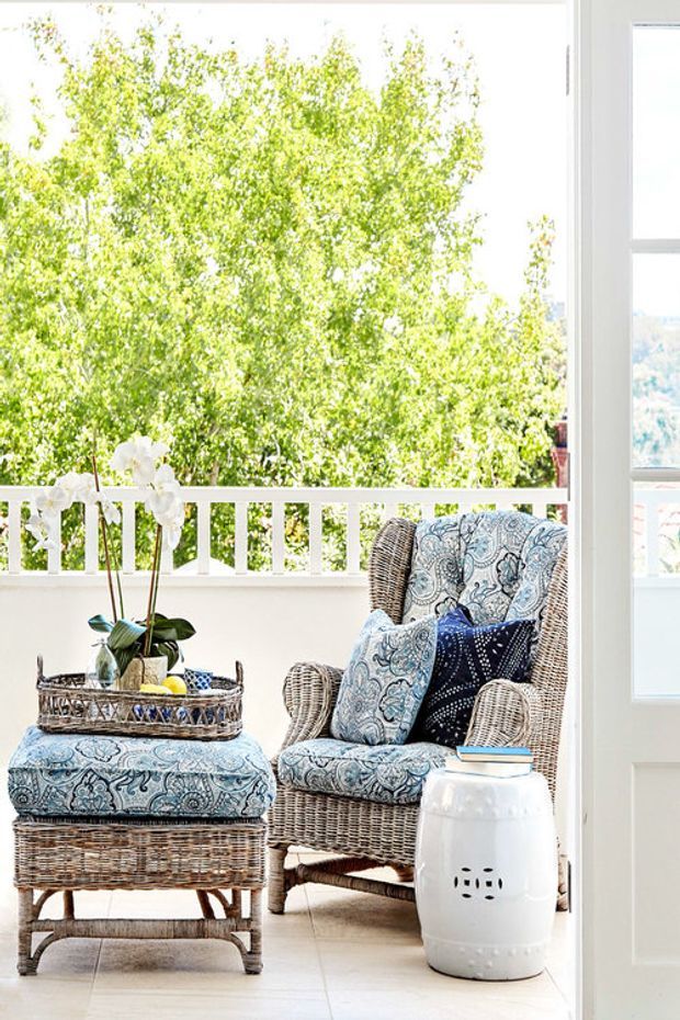 A bright, inviting outdoor veranda or enclosed balcony furnished with wicker seating and a matching ottoman upholstered in blue-and-white patterned fabric. A white balustrade, pale tiled floor, potted orchid, side table, and white ceramic garden stool frame the leafy outlook through large white-framed glazing.