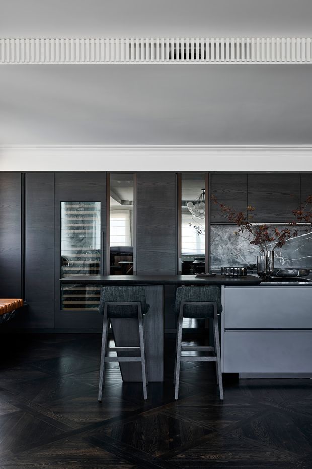 A moody, highly refined kitchen with dark timber cabinetry, a central island with two bar stools, integrated wine storage, mirrored cabinet panels, and a pale stone or marble splashback. The room features a decorative white ceiling cornice and linear ceiling vent, while the dark patterned timber floor adds depth and drama. The overall composition is sophisticated and monochromatic with subtle metallic and natural branch accents.