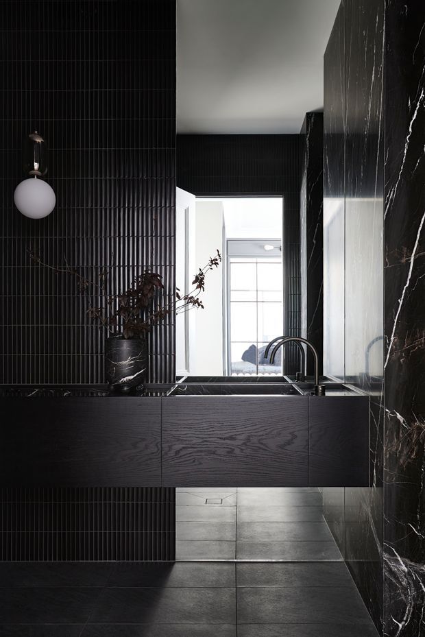 A dramatic, monochromatic kitchen or kitchenette with a black timber-grain island and integrated dark sink, black curved tap, black vertically fluted wall surfaces, dark stone flooring, and a striking black marble wall with white veining. A round wall light, dark decorative vase, and large mirror or reflective opening add contrast, while bright glazing is visible beyond.