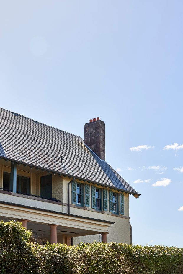 Exterior architectural photograph of a traditional rendered house with a steep grey tiled pitched roof, prominent stone chimney, timber-framed windows with green louvred shutters, and a covered veranda supported by classical columns. A clipped hedge occupies the foreground beneath a clear blue sky.