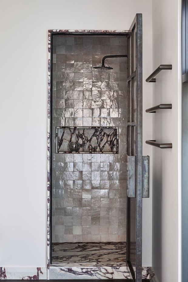 A compact, luxurious walk-in shower with reflective handmade square wall tiles in muted grey and taupe tones. The shower features a dark metal rain shower, a marble-lined recessed niche, richly veined marble flooring and threshold, and a framed glass door with dark metal hardware. A slim heated towel rail is visible on the adjacent wall.