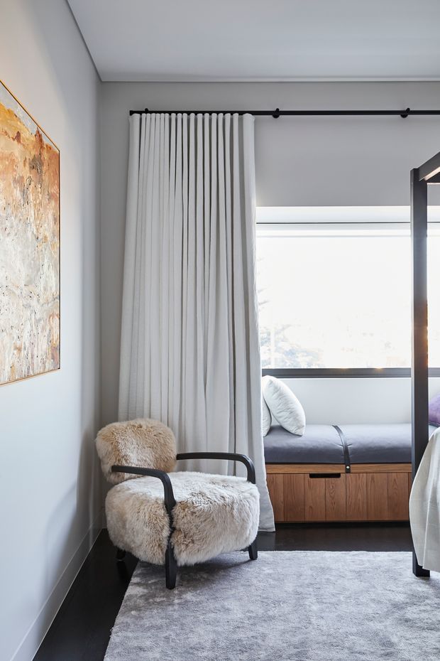 A softly lit contemporary bedroom corner featuring a black-framed armchair upholstered in cream shearling, a pale grey rug, and full-height white curtains suspended from a black ceiling track. Behind the curtains is a built-in window seat with grey cushions and warm timber cabinetry. The room has pale grey walls, a minimalist ceiling, dark flooring, and an abstract artwork on the wall.