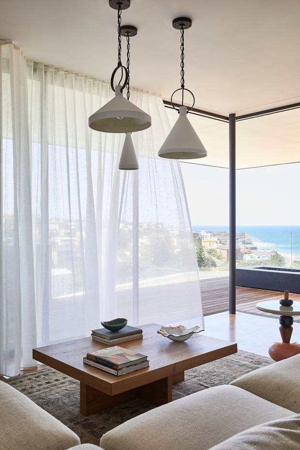 A bright coastal living room with floor-to-ceiling sheer curtains, expansive glazing, and a view toward the ocean. The room features a low rectangular timber coffee table, neutral upholstered seating, a textured rug, sculptural decorative objects, and three distinctive pendant lights suspended from the ceiling. A timber outdoor deck and dark-framed glazing extend toward the sea.