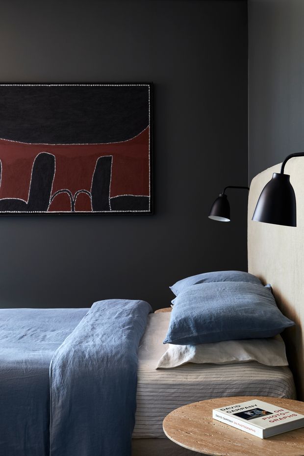 A moody, refined bedroom with a dark charcoal wall, a large framed abstract artwork, and a pale upholstered headboard. The bed is dressed in layered blue-grey linen bedding with neutral striped sheets, flanked by black wall-mounted lamps. A round natural timber side table with design books sits in the foreground.