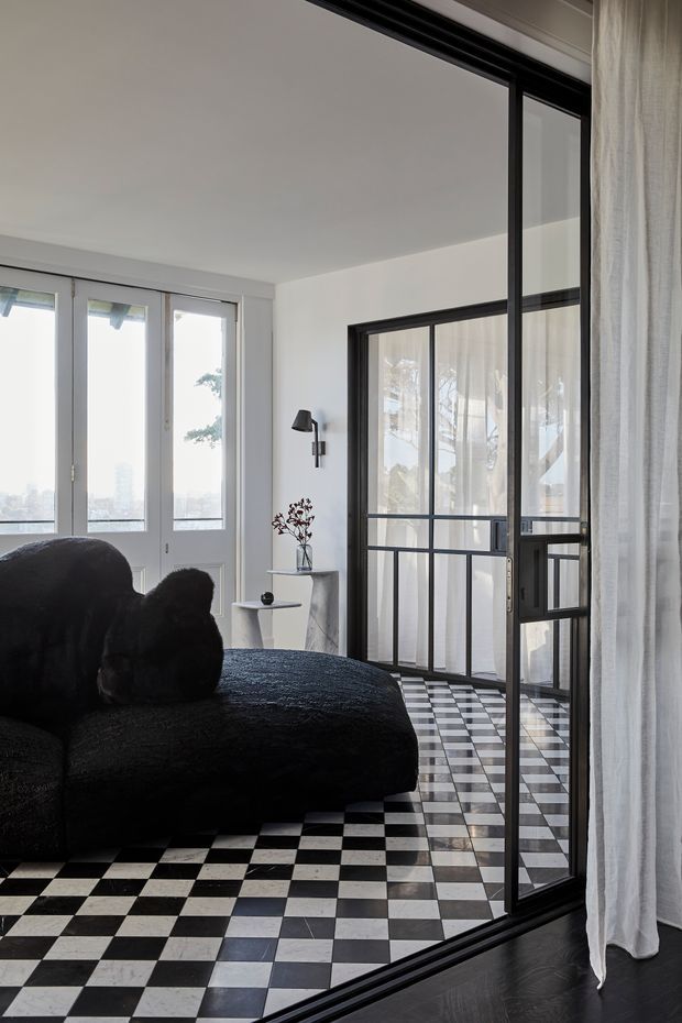 A refined contemporary living room with a dramatic black boucle sofa, black-and-white checkerboard flooring, white walls, and expansive black-framed glass partitions. Sheer curtains soften the glazing, while a small side table, wall sconce, and minimalist vase add restrained detail. Daylight enters through tall windows and doors, with a faint city view beyond.