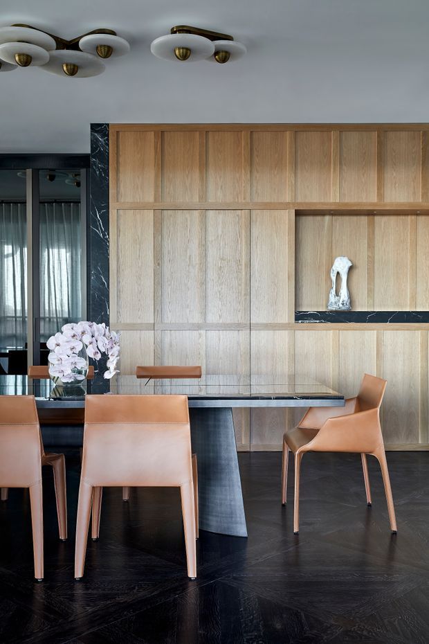A refined dining area with a dark herringbone timber floor, full-height light timber wall panelling and integrated display shelving accented with dark veined stone. A dining table with a dark stone or glass top is surrounded by tan leather dining chairs. A black-framed glazed opening with sheer curtains is visible to the left, and sculptural white-and-brass ceiling lights add a decorative focal point. The palette is warm timber, caramel leather, black and marble-like dark stone.