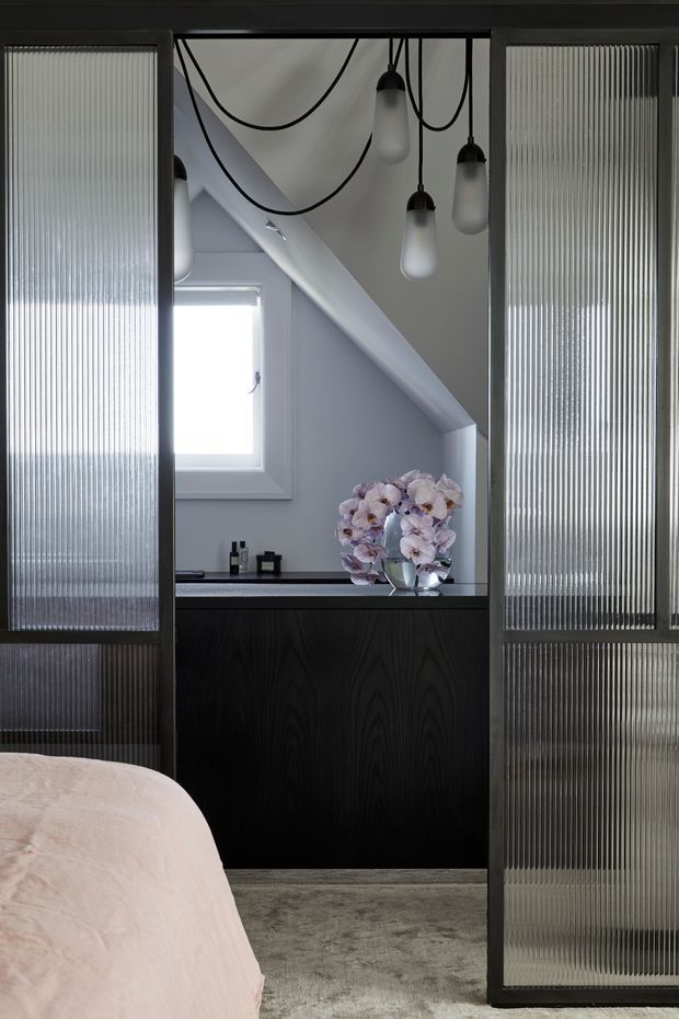 A refined bedroom interior viewed toward a compact ensuite or vanity area framed by dark metal and ribbed translucent glass sliding panels. The space features a dark timber vanity with a pale stone countertop, white walls, a small window beneath a sloping ceiling, and sculptural pendant lights with exposed black cords. A bed with pale blush bedding and an orchid arrangement is visible in the foreground, creating a moody, elegant atmosphere.