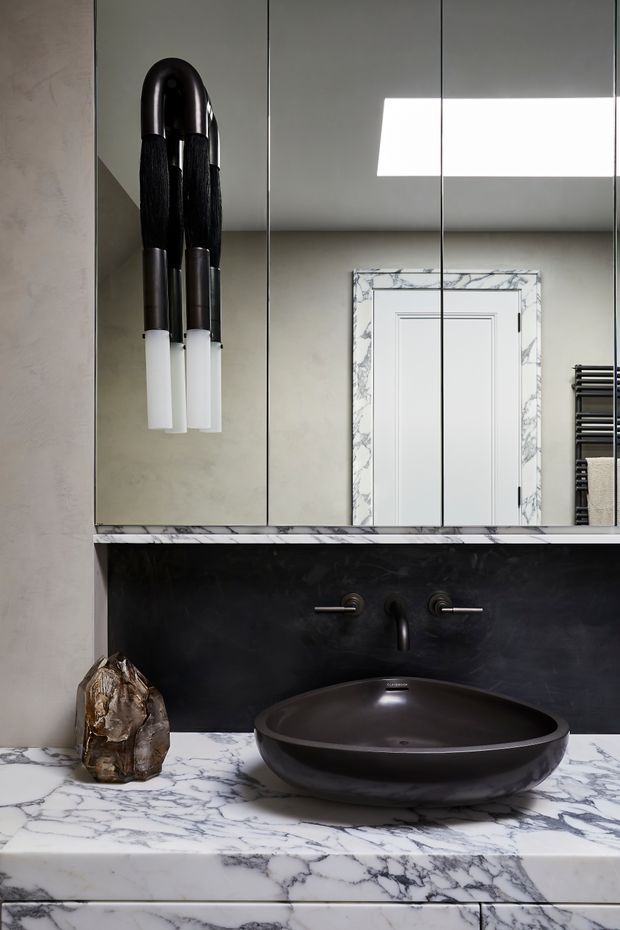 Close-up view of a refined bathroom vanity featuring a black vessel basin, wall-mounted black tapware, a dark wall backsplash, and a white marble vanity top with dramatic grey veining. Mirrored cabinetry reflects a white paneled door framed in matching marble, while a sculptural black-and-white wall light and heated towel rail add strong decorative and functional details.