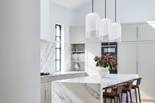 Bright, refined contemporary kitchen with a large white marble waterfall island, integrated pale cabinetry, built-in oven, marble splashback, and a trio of white glass pendant lights. A row of timber-framed bar stools lines the island, while a vase of flowers provides a small decorative accent.
