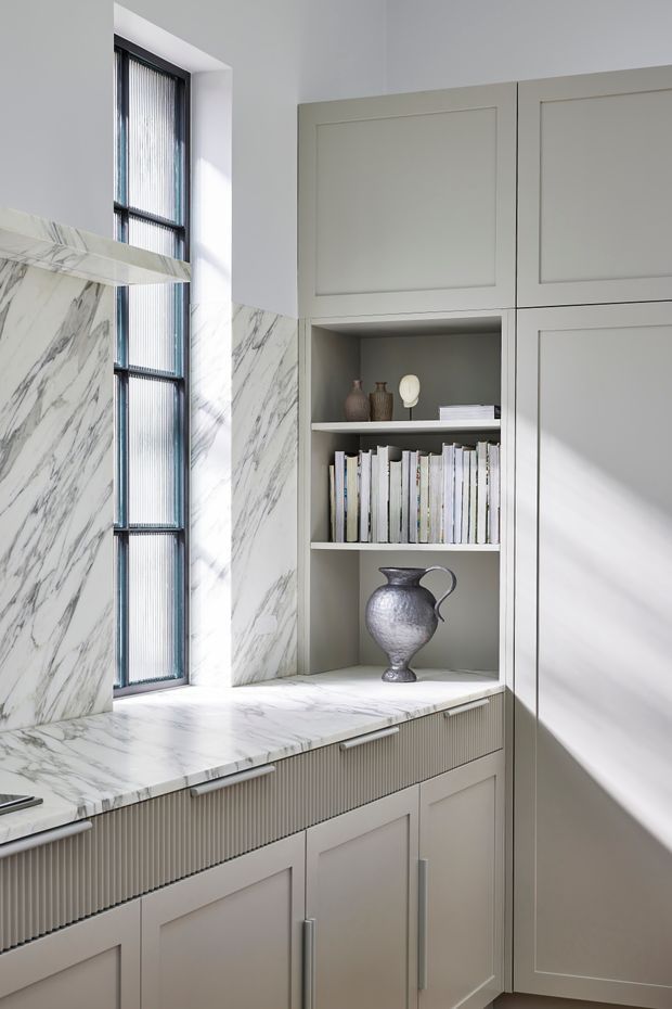 A bright, refined built-in joinery area with pale grey painted cabinetry, a white marble window bench and backsplash, open shelving displaying books and decorative vessels, and a tall black-framed ribbed-glass window. The cabinetry combines recessed-panel doors with a fluted drawer front, creating a tailored contemporary-classic look.