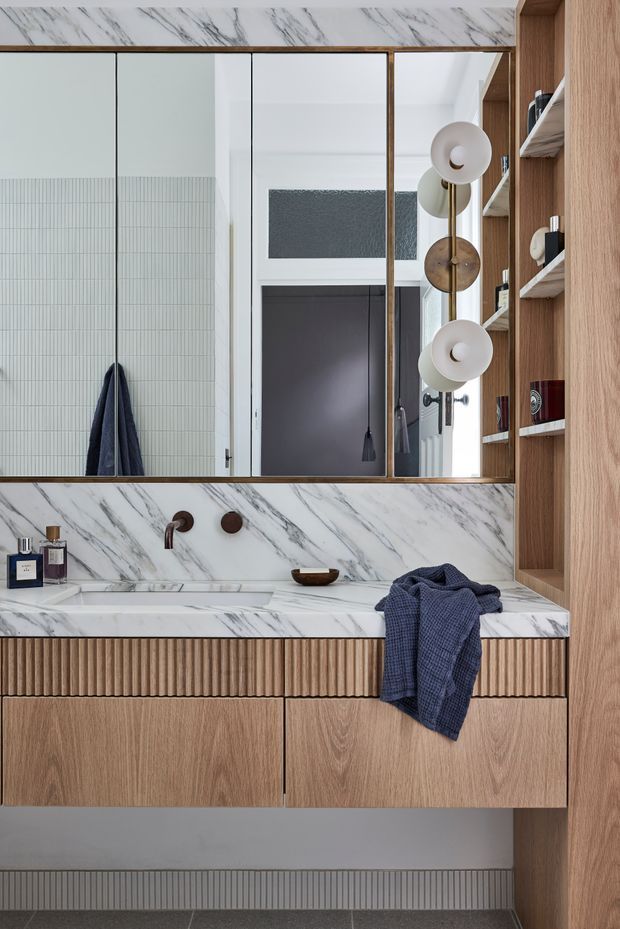 A refined contemporary bathroom vanity featuring a white marble-look benchtop and splashback with grey veining, warm timber cabinetry, integrated fluted drawer detailing, open timber shelving, bronze wall-mounted tapware, and a large mirrored cabinet. A sculptural globe light fixture and dark blue towel add contrast.