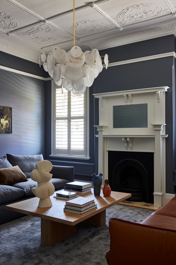 A refined living room with deep navy walls, ornate white plaster ceiling panels and cornices, a classical decorative fireplace, plantation shutters, and a sculptural white glass pendant light. The room is furnished with a dark blue sectional sofa, cognac leather seating, a timber coffee table, stacked books, sculptural decor, and a grey rug. The palette is dark and sophisticated with strong heritage detailing balanced by contemporary furniture.