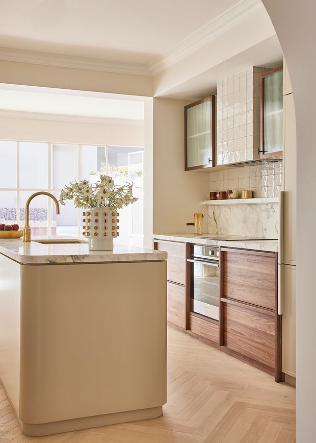 Bright, refined kitchen featuring a rounded cream island with a marble benchtop and brass gooseneck tap. The kitchen has warm timber cabinetry, integrated appliances, a marble backsplash and shelf, glazed upper cabinets with fluted glass, and a pale herringbone timber floor. Decorative cornices and arched openings add traditional detailing, while large windows bring in abundant natural light.