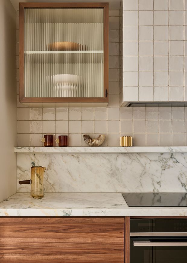 Close-up view of a refined kitchen wall and benchtop. The composition features warm timber cabinetry, a veined marble worktop and backsplash, pale square tiled walls, a black induction cooktop and built-in oven, a framed ribbed-glass cabinet, and subtle brass and ceramic accessories. The palette is warm, earthy, and luxurious with strong material contrast.