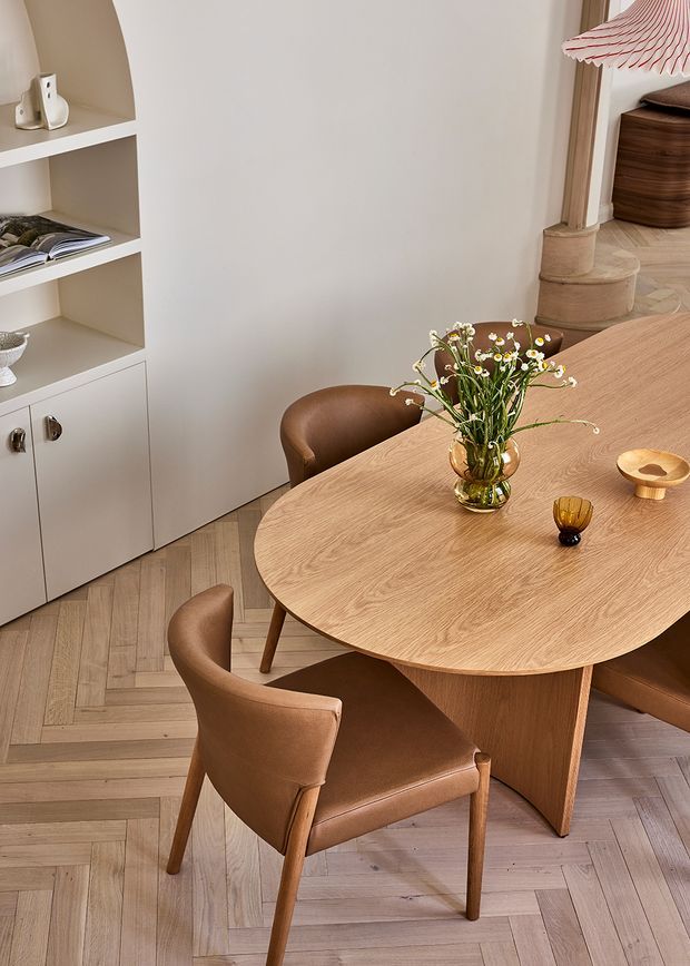 Warm, refined dining area with a large rounded timber dining table, upholstered brown leather dining chairs, and a small floral arrangement. The room features pale timber parquet flooring, built-in cream shelving and cabinetry, sculptural arched detailing, and a partially visible adjoining space with a column and wood furnishings.