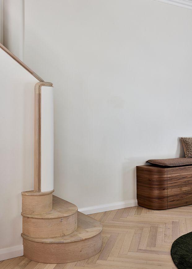 A refined interior corner featuring a curved timber staircase with a slender light-toned balustrade, pale herringbone timber flooring, and a dark walnut curved sideboard topped with upholstered seating. The walls and trim are soft white, creating a calm, understated composition with sculptural rounded forms and warm natural wood.
