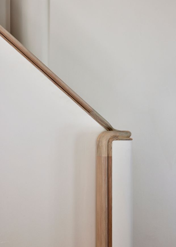 Close-up architectural detail of a warm timber handrail and vertical stair or balustrade element meeting at a carefully rounded junction against a smooth off-white plaster wall. The image is minimal, softly lit, and focused on refined joinery and natural wood grain.