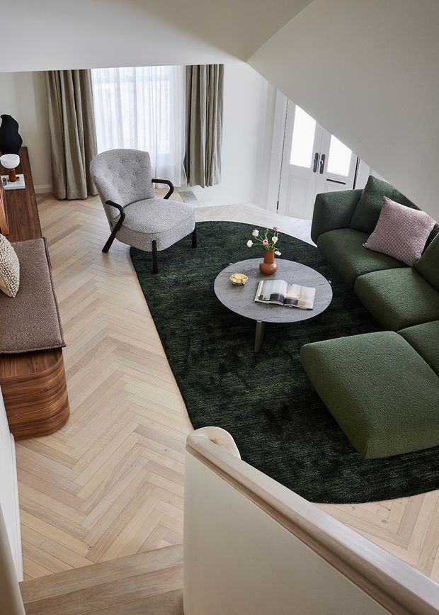 A refined living room viewed from a stair landing, furnished with a deep green sectional sofa, textured grey armchair, round dark coffee table, and dark green area rug. The room features pale herringbone timber flooring, white walls and cabinetry, full-height curtains, and abundant natural light. A curved stair balustrade frames the foreground.