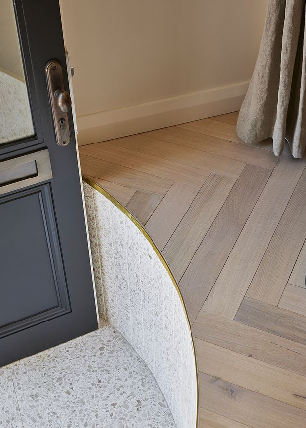 Close-up architectural detail of a threshold between an exterior terrazzo landing and an interior timber herringbone floor. A dark paneled door with a metallic lock and glazed panel opens inward, while a curved terrazzo partition or step edge with a slim brass-toned trim separates the two floor finishes. Neutral painted walls and a textured curtain are visible.