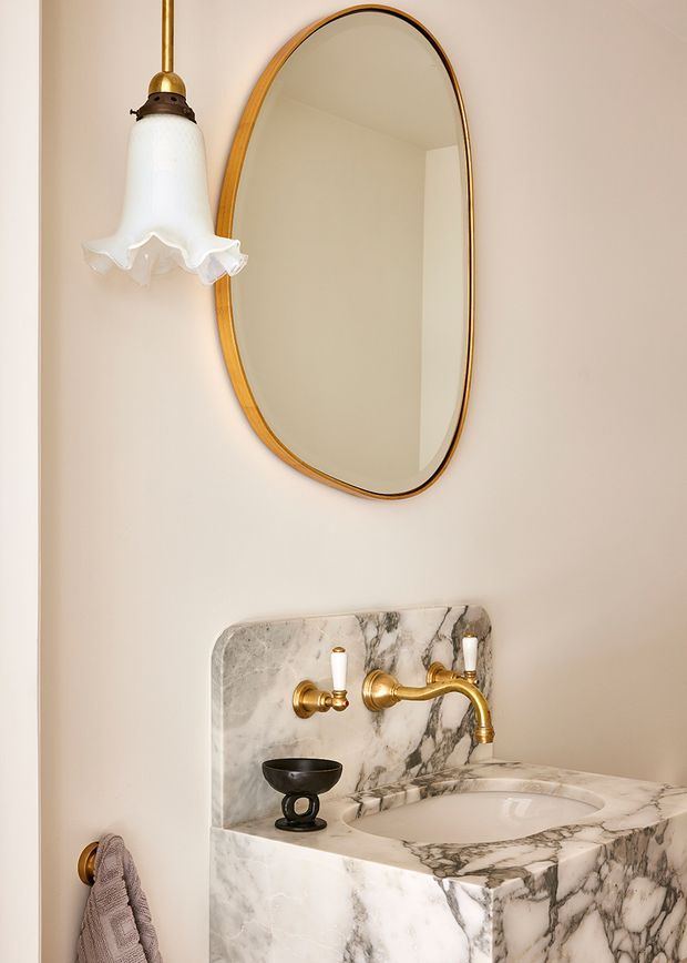 A compact, refined bathroom vanity with a sculpted white marble basin and matching marble splashback, featuring prominent grey veining. A brass wall-mounted tap set, oval brass-framed mirror, and decorative pendant light create a warm, traditional character. A small towel rail with a hand towel is visible at the side.