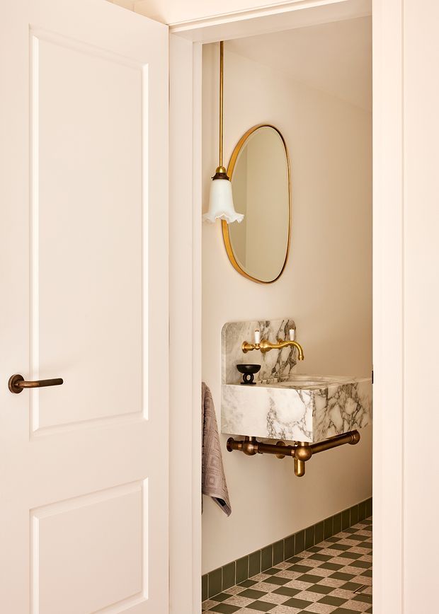A compact powder room viewed through an open white paneled door. The room features a wall-mounted marble basin with dramatic veining, antique brass plumbing and tapware, an oval brass-framed mirror, a decorative wall sconce, warm beige walls, and a green-and-cream checkerboard tile floor with matching dark green skirting tiles.