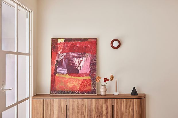 A warmly styled interior vignette featuring a low timber sideboard with vertical cabinet doors, topped by a large abstract red and pink artwork, ceramic vessels, candles, and a dark sculptural object. A tall glazed door or window with a light-coloured frame is visible on the left, while a small circular wall mirror adds a decorative accent. The composition has a refined, contemporary feel with natural timber and muted neutral walls.