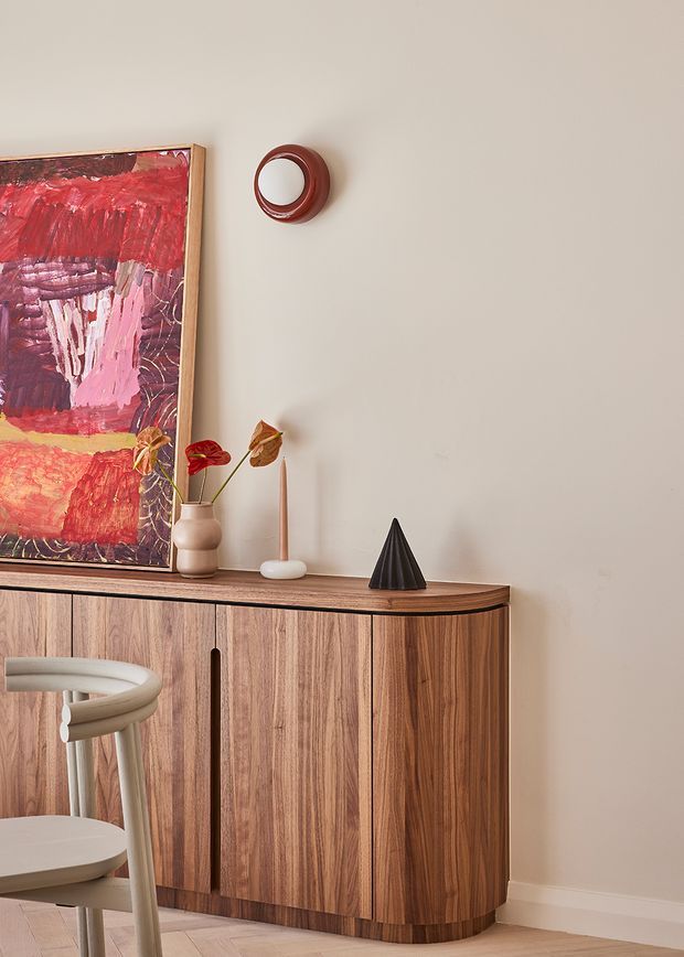 A warm, curated interior vignette featuring a mid-century-style timber sideboard with rounded corners and vertical wood grain, displayed against a soft neutral wall. An abstract red and pink artwork, ceramic vase with dried stems, candle, sculptural black ornament, and circular wall object add decorative interest. A light upholstered dining chair is partially visible in the foreground, with pale timber flooring below.