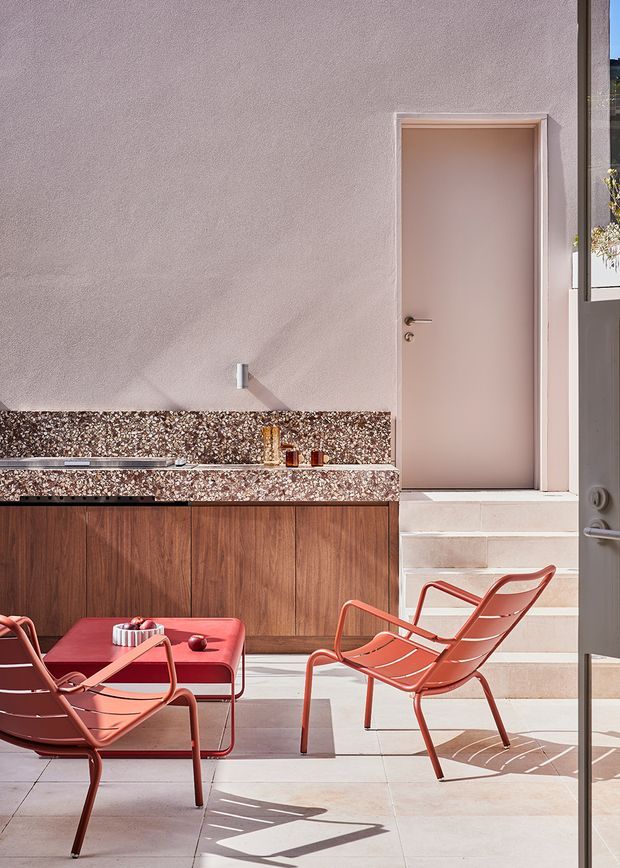 A sunlit outdoor courtyard or terrace with a compact built-in outdoor kitchen along a textured stucco wall. The kitchen features warm timber cabinetry, a terrazzo-like stone benchtop with integrated sink, and a simple wall tap. Coral-red metal lounge chairs and a matching low table sit on pale tiled paving beside a short flight of steps leading to a plain door. The scene has a warm, refined mid-century-inspired aesthetic with strong geometric shadows.