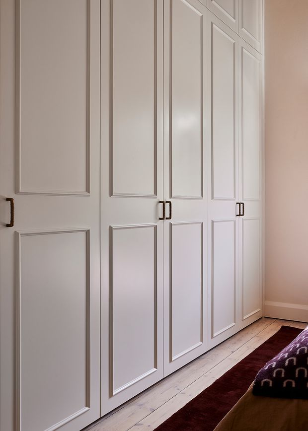A close view of tall, built-in white paneled wardrobe cabinetry with dark metal pull handles in a softly lit bedroom. Pale timber flooring, a burgundy runner rug, and the edge of a bed are visible, creating a refined traditional interior.