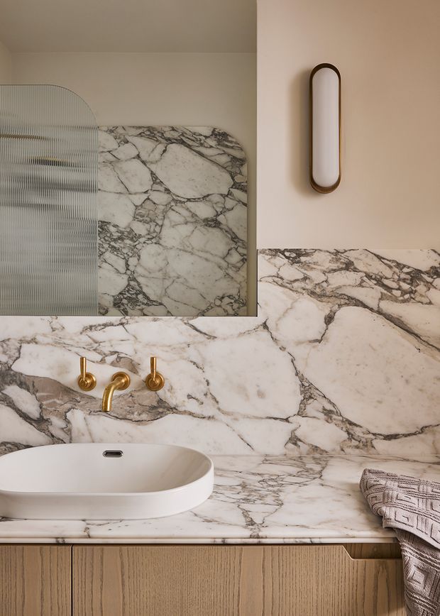 Close-up view of a refined bathroom vanity featuring a white vessel basin, a wall-mounted brushed brass faucet set, and dramatic white-and-grey veined marble extending across the benchtop and backsplash. The vanity has vertically fluted timber cabinetry, a rounded-edge mirror, and a slim oval wall light with a dark bronze or brass frame. Neutral, warm, high-end material palette.