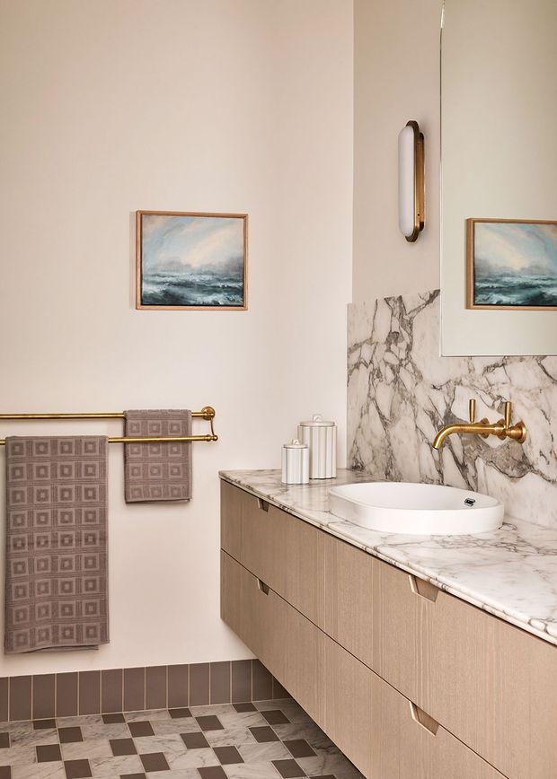 A refined contemporary bathroom featuring a floating light-wood vanity with integrated drawers, a white vessel basin, and a polished brass wall-mounted tap. The vanity is topped with heavily veined marble extending into a matching backsplash. A large mirrored cabinet, brass towel rail, patterned neutral towels, wall sconce, framed seascape artwork, and a contrasting checkerboard floor complete the warm, elegant scheme.