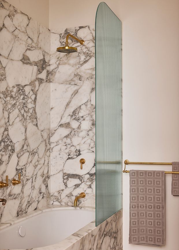 A luxurious bathroom featuring a freestanding or inset white bathtub surrounded by dramatic veined marble wall and bath-panel cladding. A tall curved-edge fluted glass shower screen separates the bath area, while polished brass fixtures, towel rails, and taps add warmth. Neutral plaster walls and patterned taupe towels complete the refined interior.