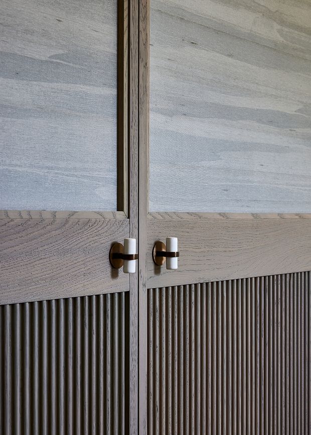 Close-up detail of bespoke timber cabinetry or wardrobe doors. The doors combine pale, heavily grained timber panels with lower vertically fluted timber panels, slim timber framing, and contrasting dark metal-and-white hardware. The image is tightly cropped with no wider room context visible.