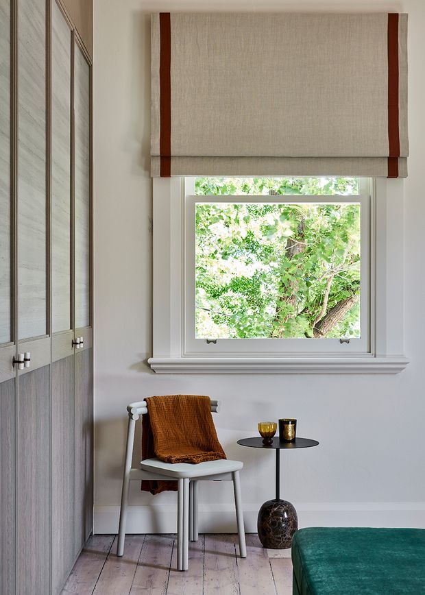 A refined bedroom corner with full-height built-in wardrobe cabinetry along the left wall, a classic white-trimmed window with a textured Roman blind, and a small white-framed chair draped in a rust-brown textile. A dark pedestal side table with two glasses sits beside the chair, while the edge of a deep green upholstered bed or bench is visible in the foreground. The room combines painted white walls and trim with pale timber floorboards and muted, warm furnishings.