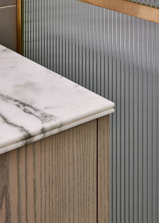 Close-up detail of a refined built-in counter or cabinet, featuring a white marble benchtop with prominent grey veining, vertically fluted timber cabinetry below, and a vertically ribbed metallic grey wall finish behind it. A warm timber trim runs along the upper edge.