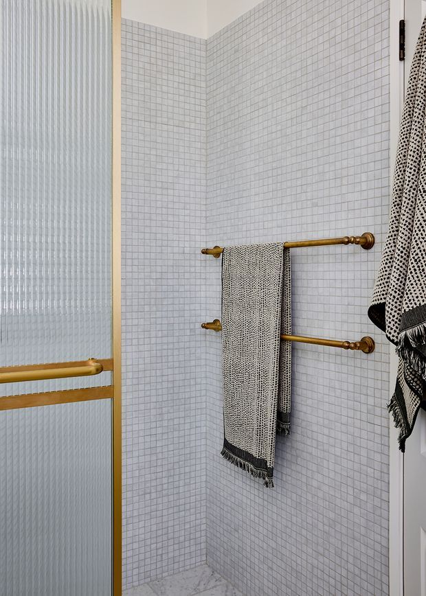 Close-up view of a compact bathroom shower area with small pale square wall tiles, a matching light stone-look floor, a textured translucent glass door in a warm brass/gold frame, and two brass towel rails holding patterned black-and-white towels. The space has a refined, contemporary look with traditional brass detailing.