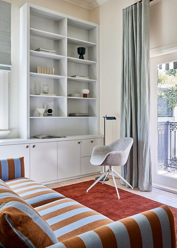 A refined sitting room with a built-in white shelving and cabinetry wall displaying books and decorative objects. The room features a striped rust, blue-grey, and cream sofa, a light upholstered lounge chair, a slim black floor lamp, a rust-red area rug, tall grey curtains, and glazed doors opening to a balcony. Traditional mouldings and a light timber floor frame the bright, tailored interior.