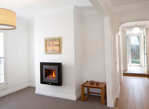 Bright traditional interior view of a hallway or transitional living area, featuring white walls with decorative cornice detailing, a built-in contemporary black fireplace, carpeted flooring in the foreground, and a timber console table. An arched opening leads to a timber-floored corridor and glazed exterior door, with abundant natural light and classic white mouldings.