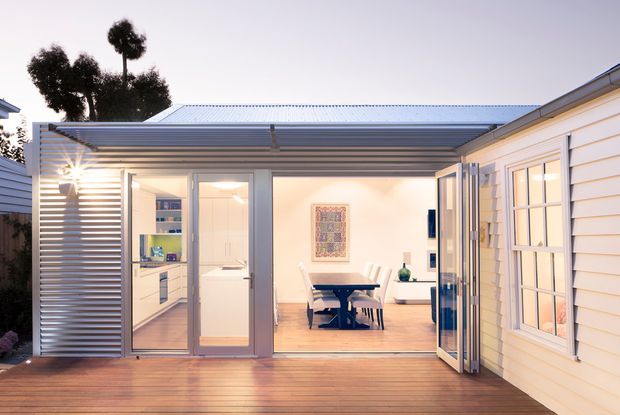 A contemporary single-storey residential extension viewed from a timber deck, with a wide glazed opening connecting the outdoor area to a bright dining and living space. The extension has a low corrugated metal roof, horizontal metal screening, aluminium-framed glazing, pale timber flooring, white cabinetry, and a white weatherboard-clad building wing. A dining table, chairs, artwork, and built-in joinery are visible inside.
