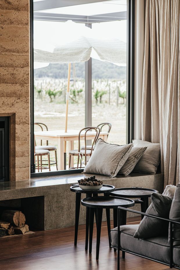 A refined living room with a stone fireplace, dark timber flooring, floor-to-ceiling glazing, and full-height neutral curtains. A grey upholstered lounge chair, layered black nesting tables, and cushions create a warm seating area beside the window. Through the glazing, an outdoor dining setting, vineyard-like planting, and hills are visible.