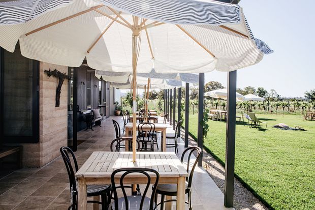 A covered outdoor dining terrace beside a dark-clad building, furnished with timber dining tables and black bentwood-style chairs beneath large striped umbrellas. Slim dark posts frame views across a manicured lawn and vineyard, creating a relaxed hospitality-style setting with strong indoor-outdoor connection.