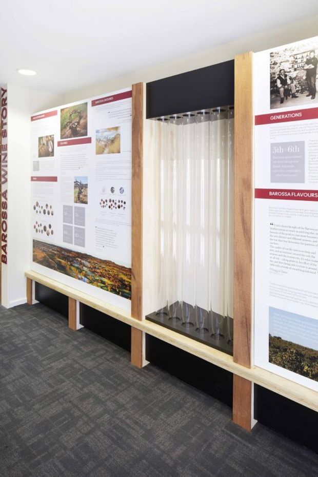 A contemporary exhibition or visitor-centre interior featuring wall-mounted interpretive panels about Barossa wine history and flavours. Timber-framed display bays with black backing surround a suspended arrangement of clear wine glasses, while grey patterned carpet covers the floor. The space is bright and educational, with integrated recessed ceiling lighting.