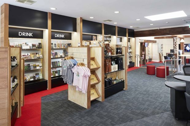 A bright, professionally fitted commercial retail and hospitality interior with wall-mounted product displays, open shelving, clothing racks, beverage merchandise, and a seating area. The space uses dark feature walls, warm timber display framing, patterned grey carpet, and bold red carpet accents. Recessed ceiling lights and a large skylight provide even illumination, while a visible welcome area and branded display zones reinforce the commercial setting.